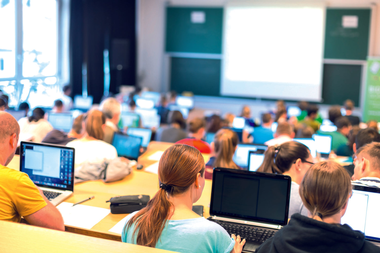 Students in classroom using laptops at desks, attending lecture with projector screen