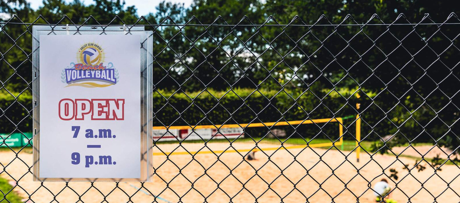 Beach volleyball court open sign on chain-link fence, hours 7am–9pm, sandy court visible behind