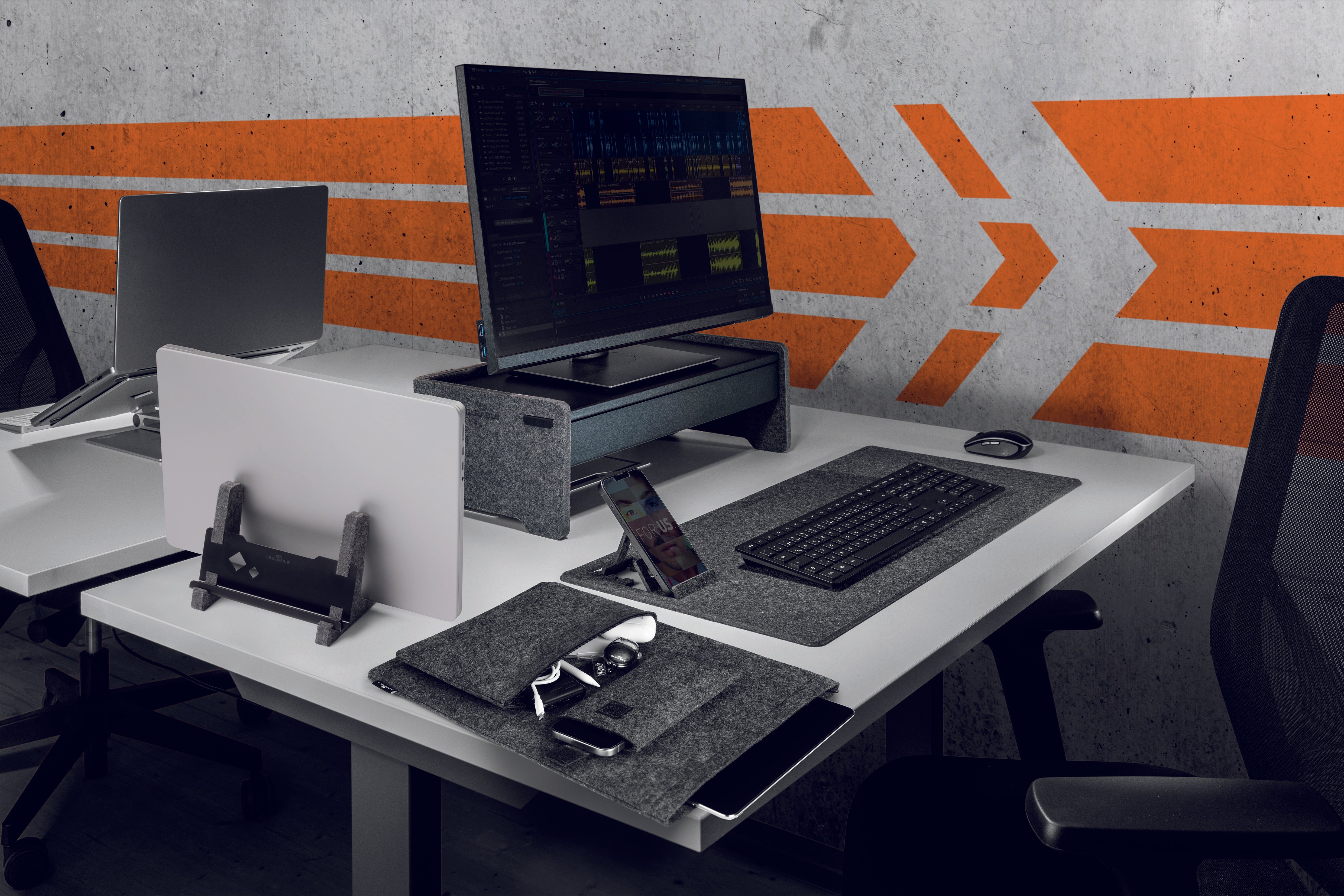 Modern office desk setup with monitor stand, laptop, keyboard, and accessories on white desk against concrete wall with orange arrows
