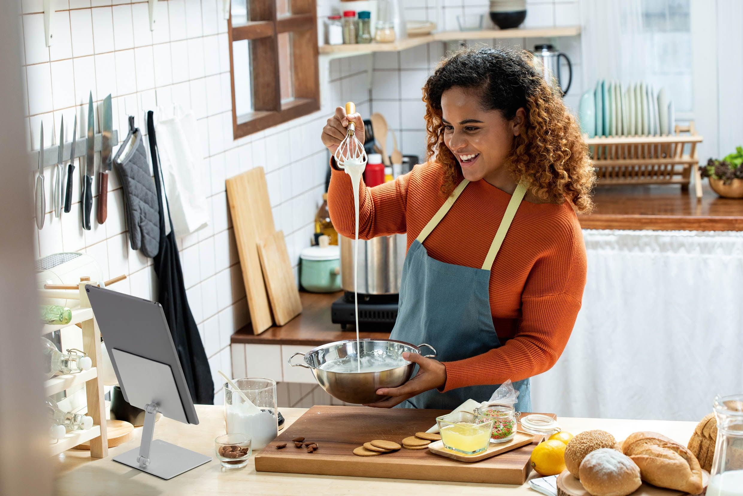 Woman baking in kitchen with tablet stand, mixing batter with whisk, fresh bread and ingredients nearby