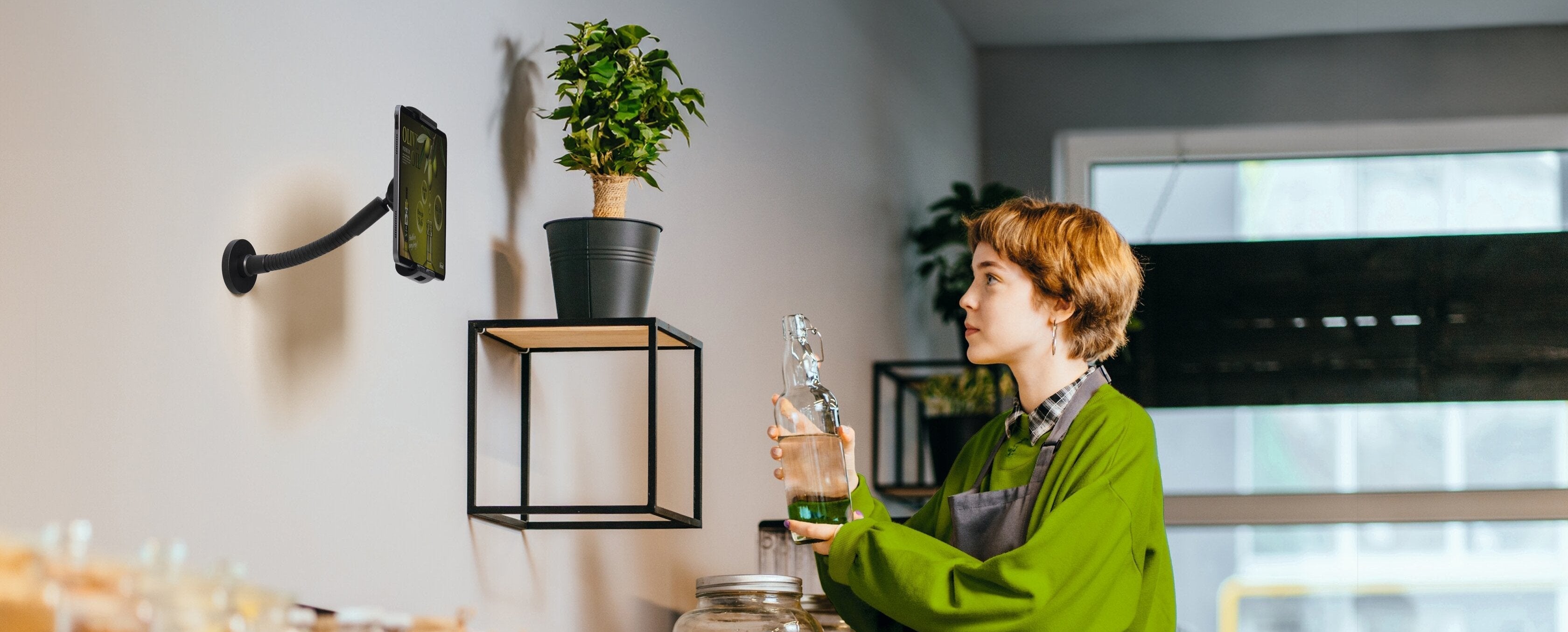 Wall-mounted tablet holder in modern kitchen with woman holding glass bottle, green plant on shelf