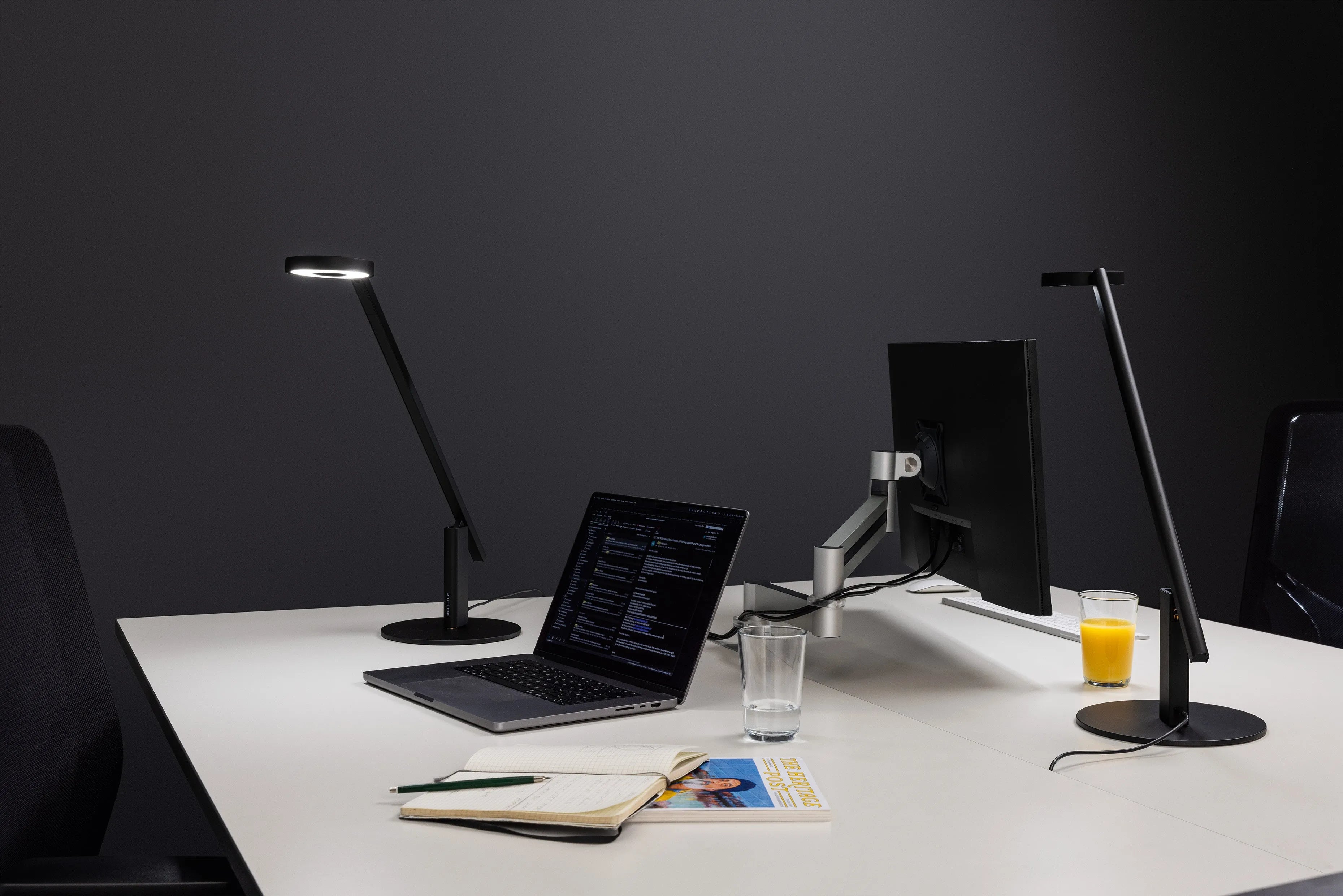 Modern office desk setup with sleek black task lamps, laptop, monitor, notebook, and drinks for productive workspace