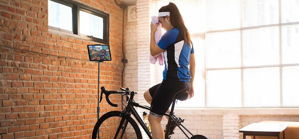 Woman on stationary bike using tablet for workout, wiping sweat with towel in bright room