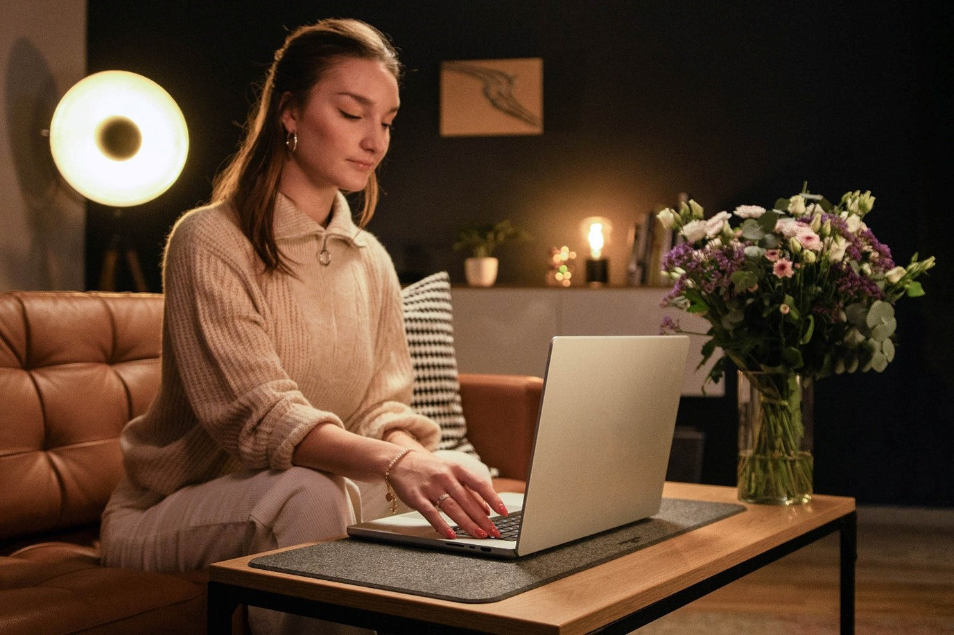 Woman typing on laptop at cozy home office desk with flowers and warm lighting